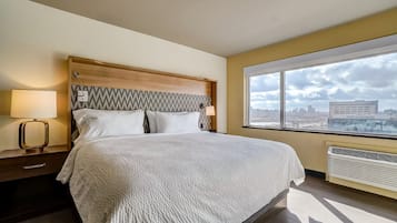 A hotel room with a large bed, a nightstand, a lamp, a window with a city view, and a wall-mounted air conditioner.