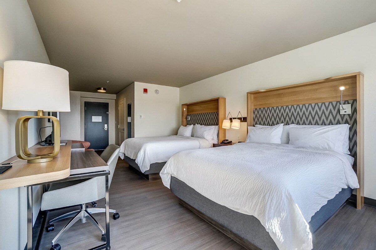 A hotel room with two beds, a desk with a lamp, and a chair.