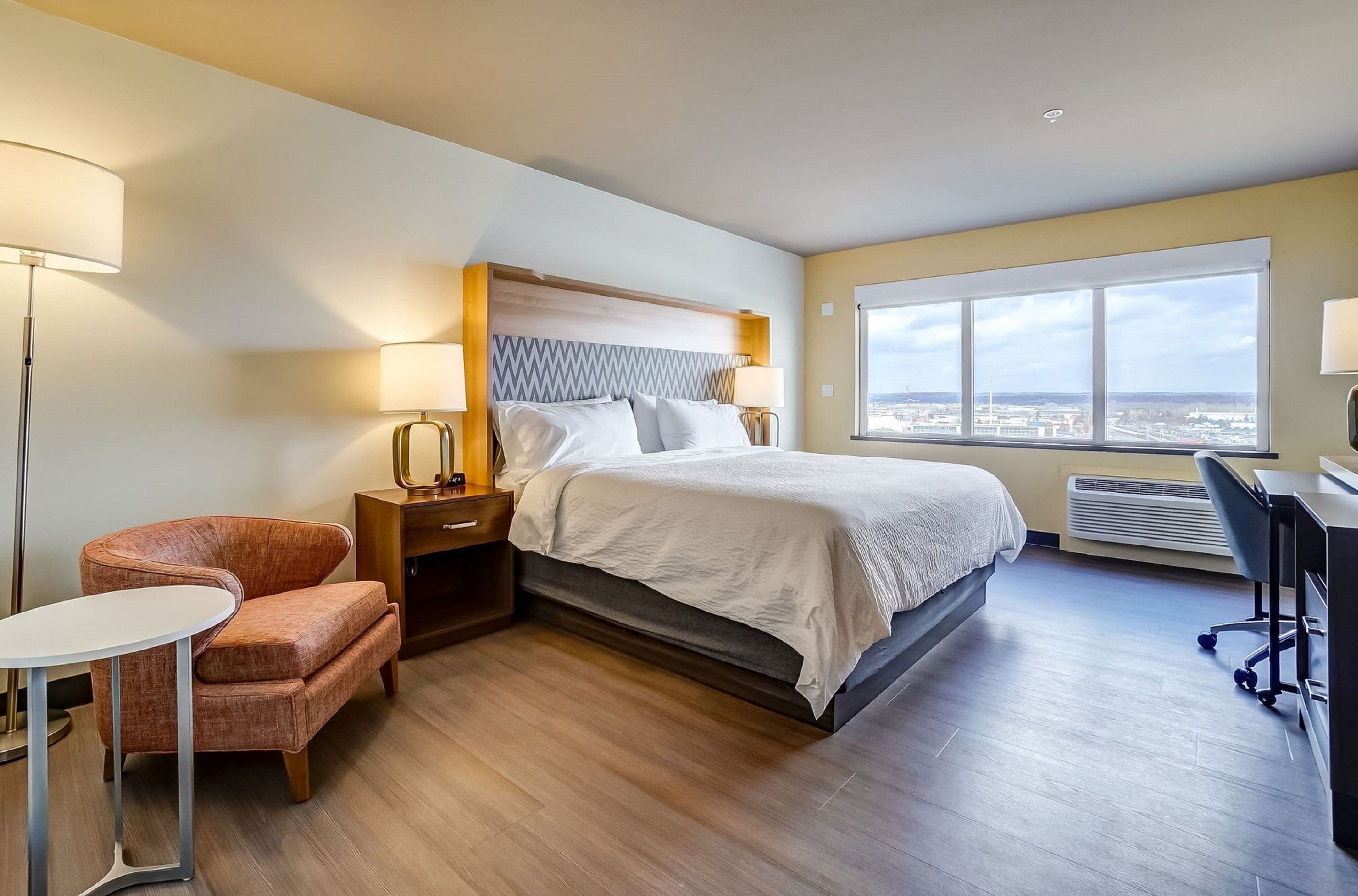 A hotel room with a bed, a desk, a chair, and a large window offering a city view.