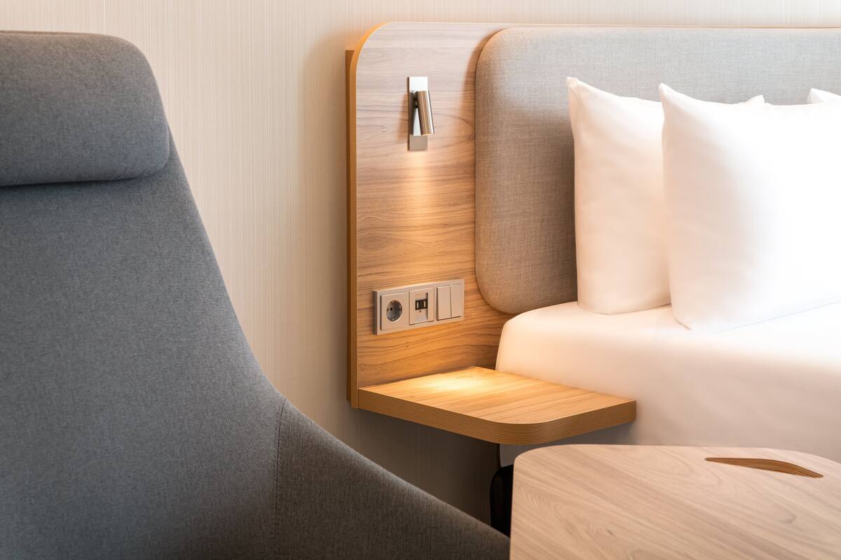 A modern hotel room with a curved grey armchair, a wooden bedside table, and a bed with white linens.
