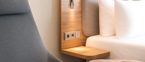 Una habitación de hotel moderna con un sillón gris curvo, una mesita de noche de madera y una cama con ropa de cama blanca.