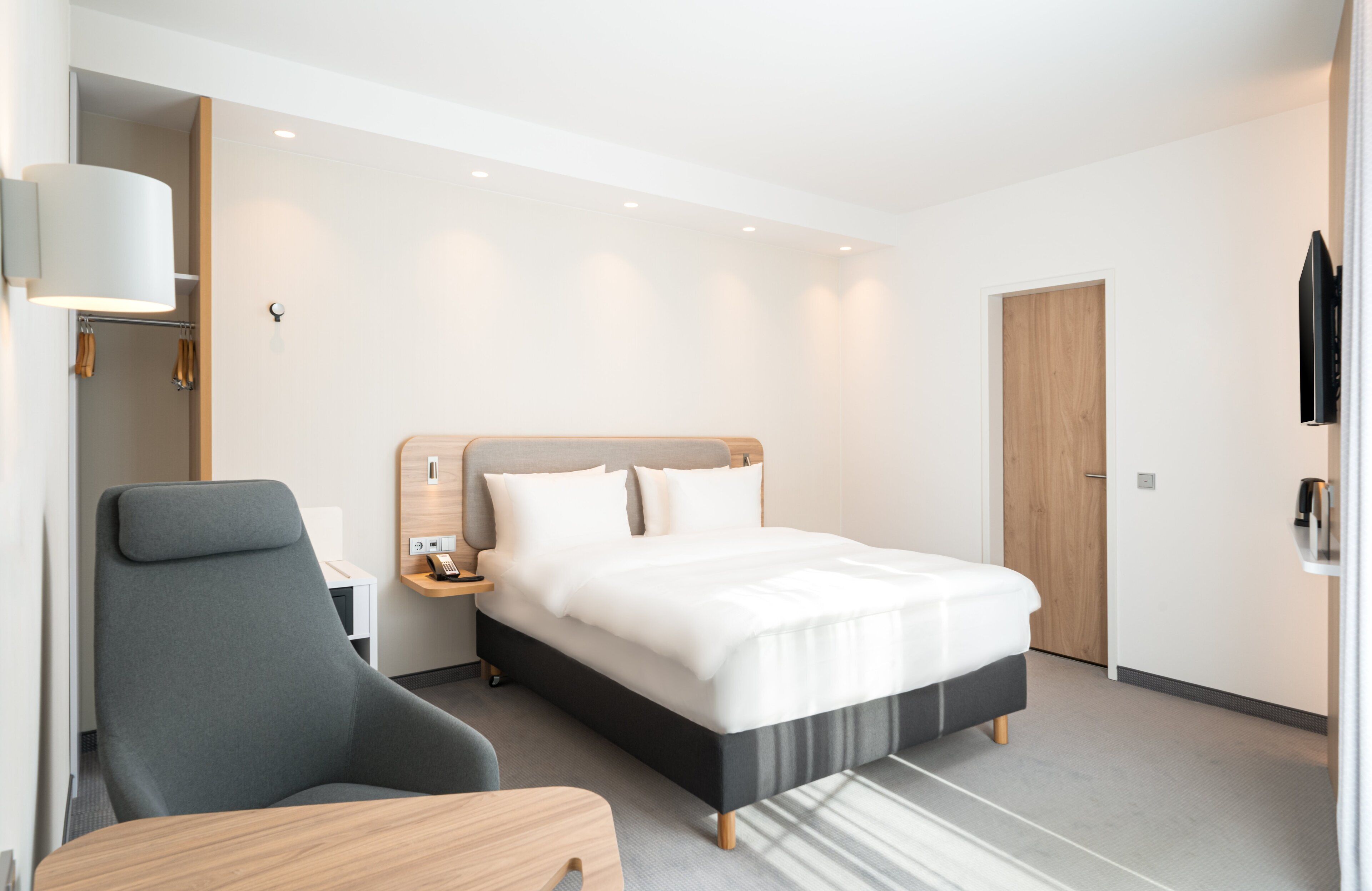 Una habitación de hotel moderna con una cama grande, un sillón gris, una mesita de noche y una puerta de madera.