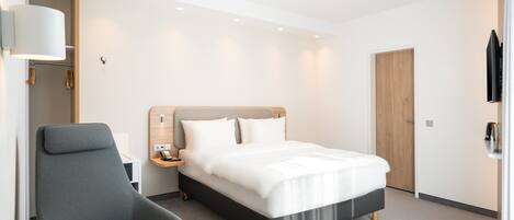 Una habitación de hotel moderna con una cama grande, un sillón gris, una mesita de noche y una puerta de madera.