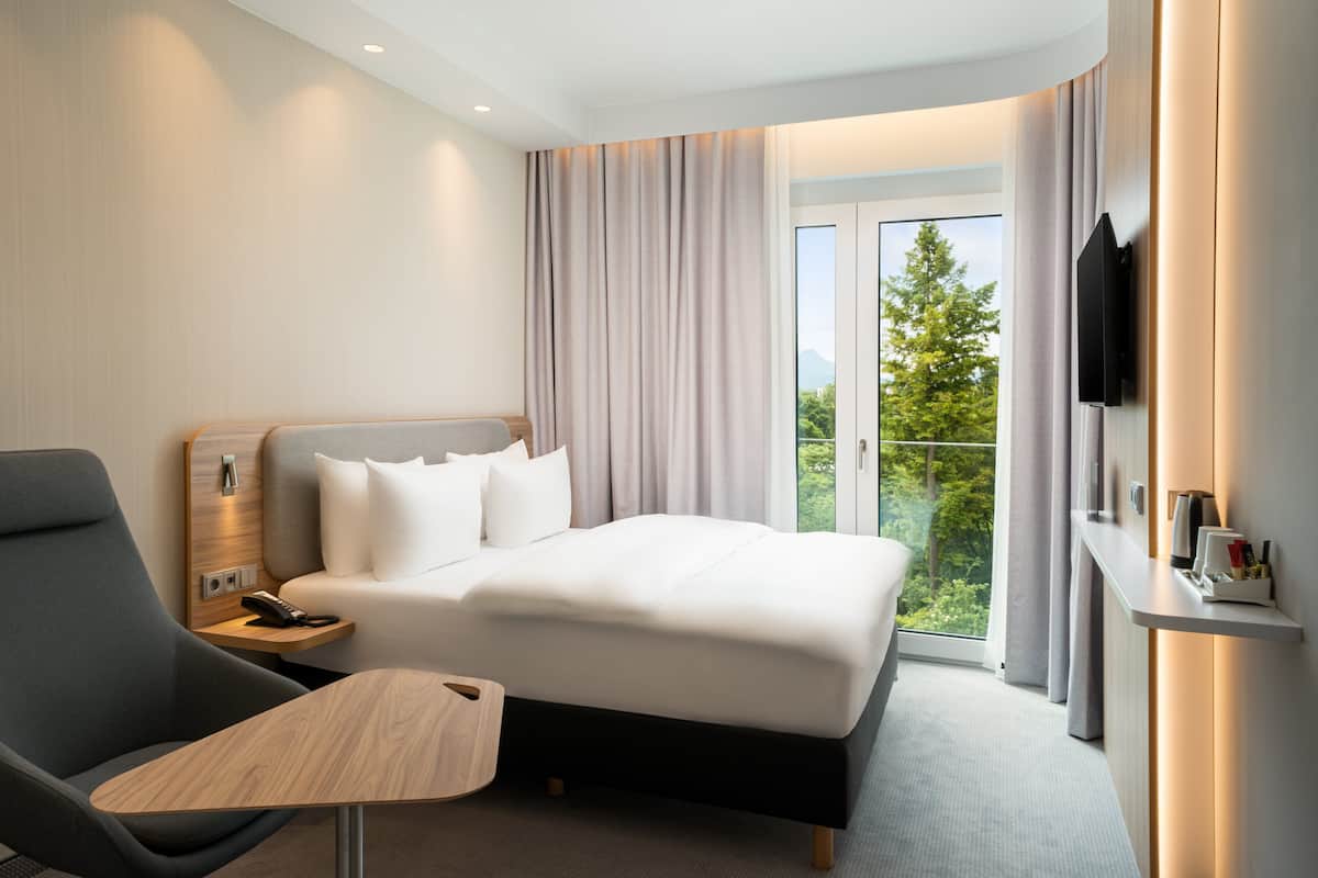 Ein Hotelzimmer mit Bett, Schreibtisch, Sessel, Fernseher und einem Balkon mit Blick auf Bäume.