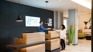 A hotel reception area with wooden reception desks, a digital screen displaying the hotel name, and directional signs for amenities.
