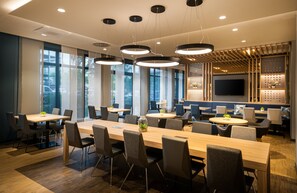 A modern conference room with a long table, multiple chairs, pendant lighting, and a large screen on the wall.