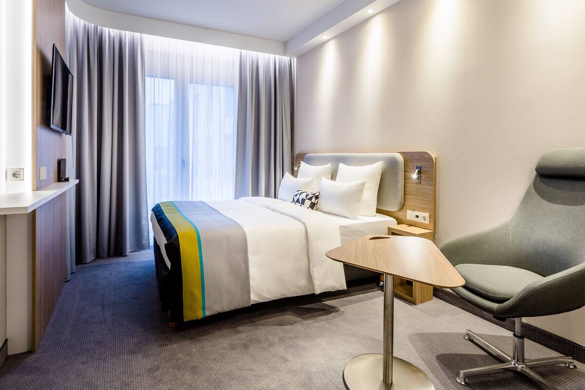 A hotel room with a bed, a TV mounted on the wall, a chair, a small table, and a window with curtains.