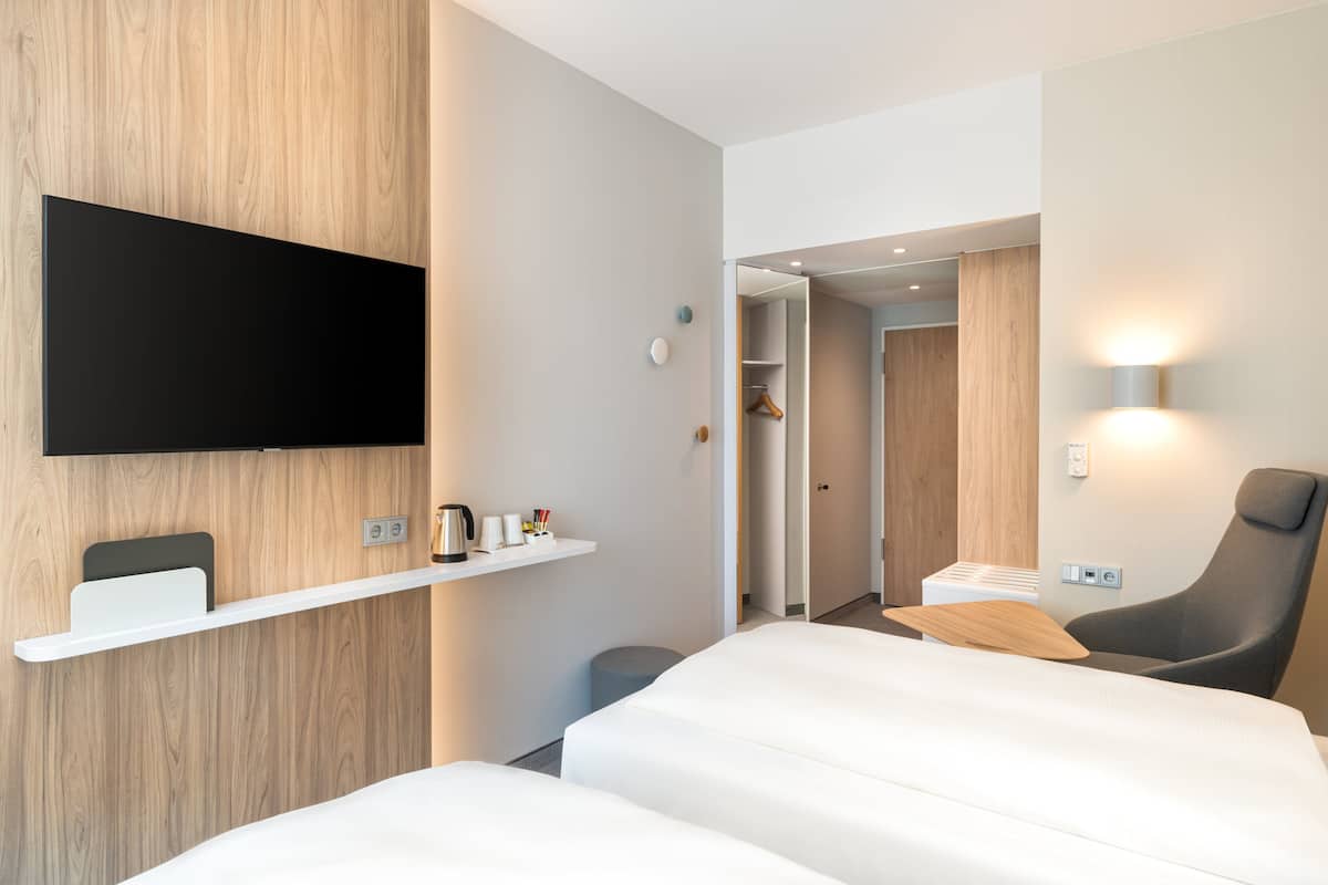 Ein modernes Hotelzimmer mit flachbildfernseher an der Wand, einem Nachttisch mit Wasserkocher, Tassen und einer kleinen Pflanze, einem großen Bett mit weißer Bettwäsche, einem grauen Sessel und einem Holzboden.