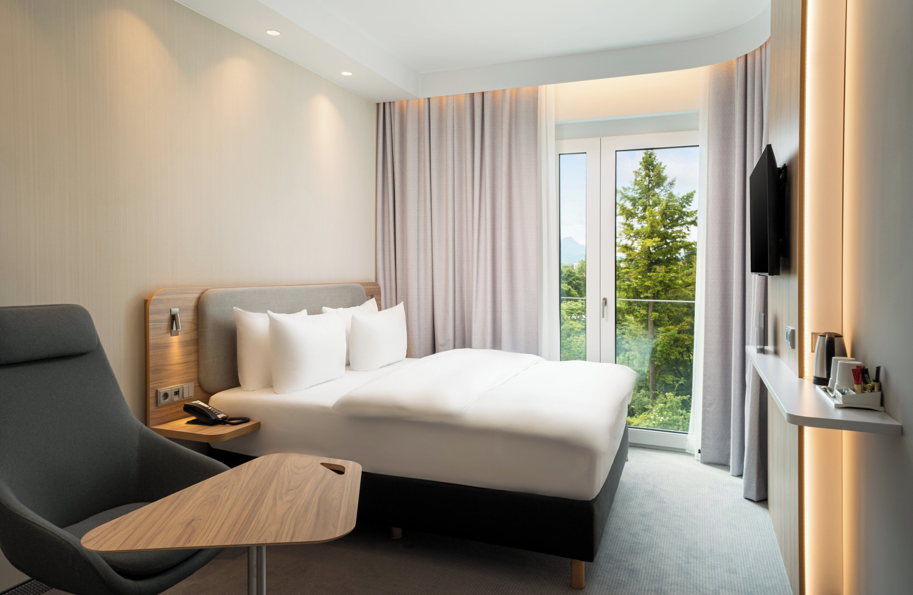 A hotel room with a bed, a desk, a chair, a TV, and a balcony with a view of trees.