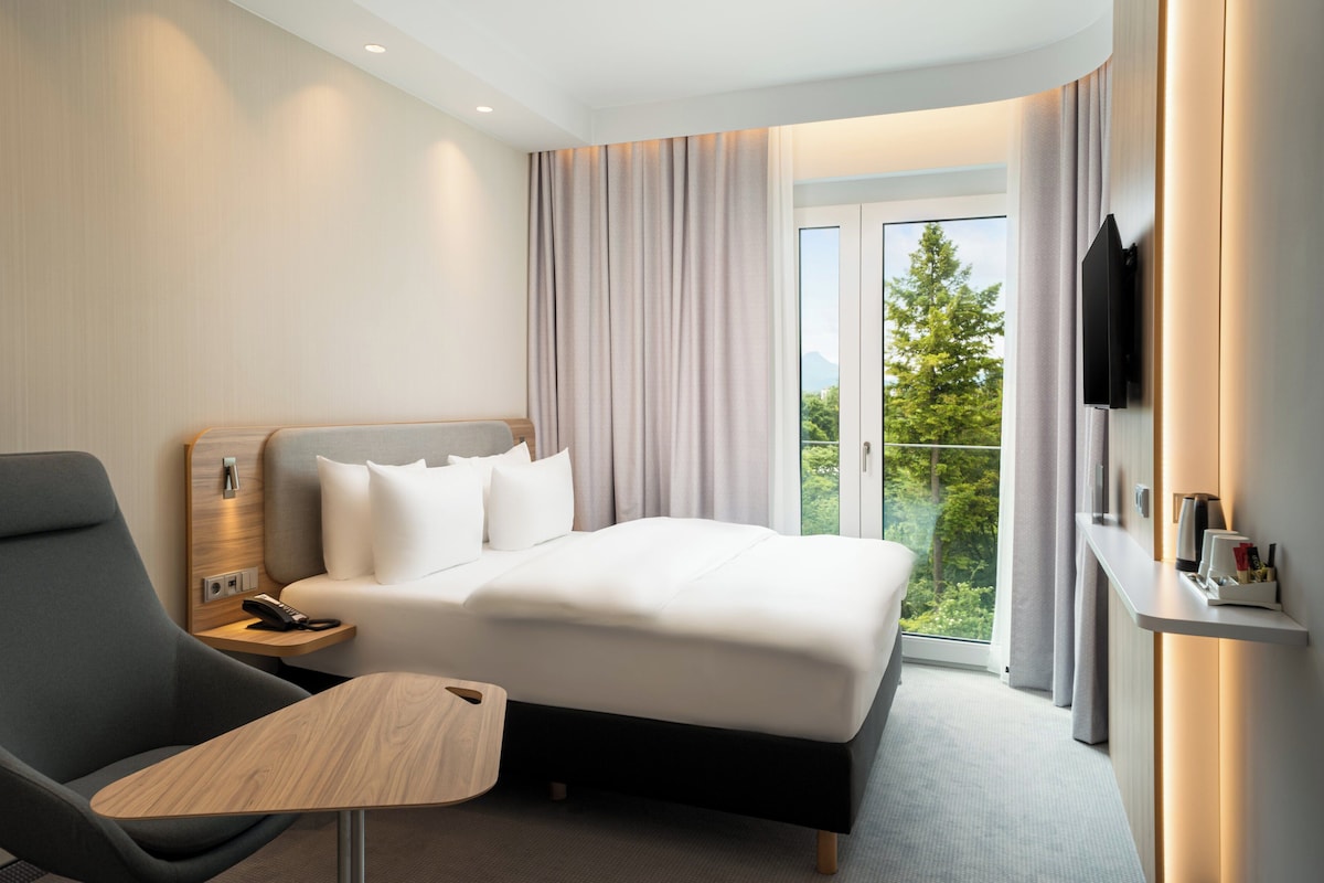 Habitación de hotel con cama, escritorio, silla, televisor y balcón con vistas a árboles.