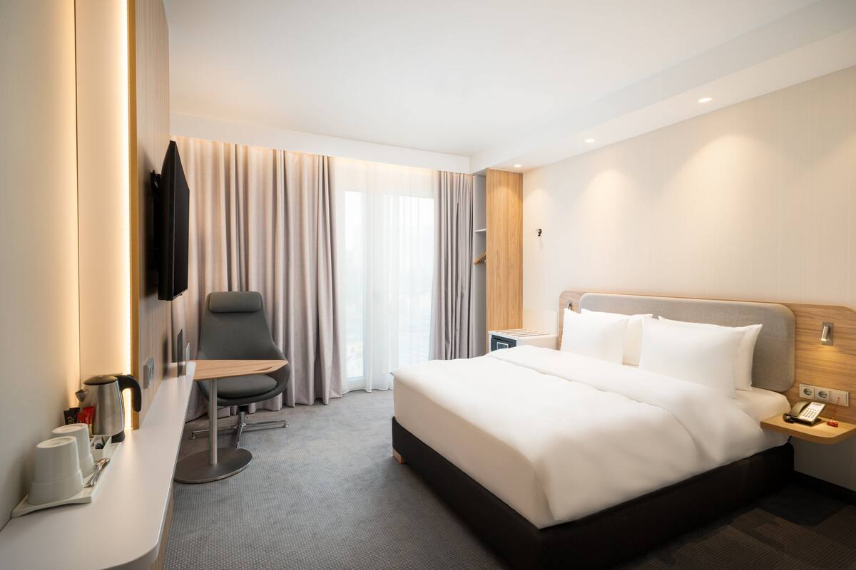 A modern hotel room with a large bed, a desk with a chair, a TV, and a window with curtains.