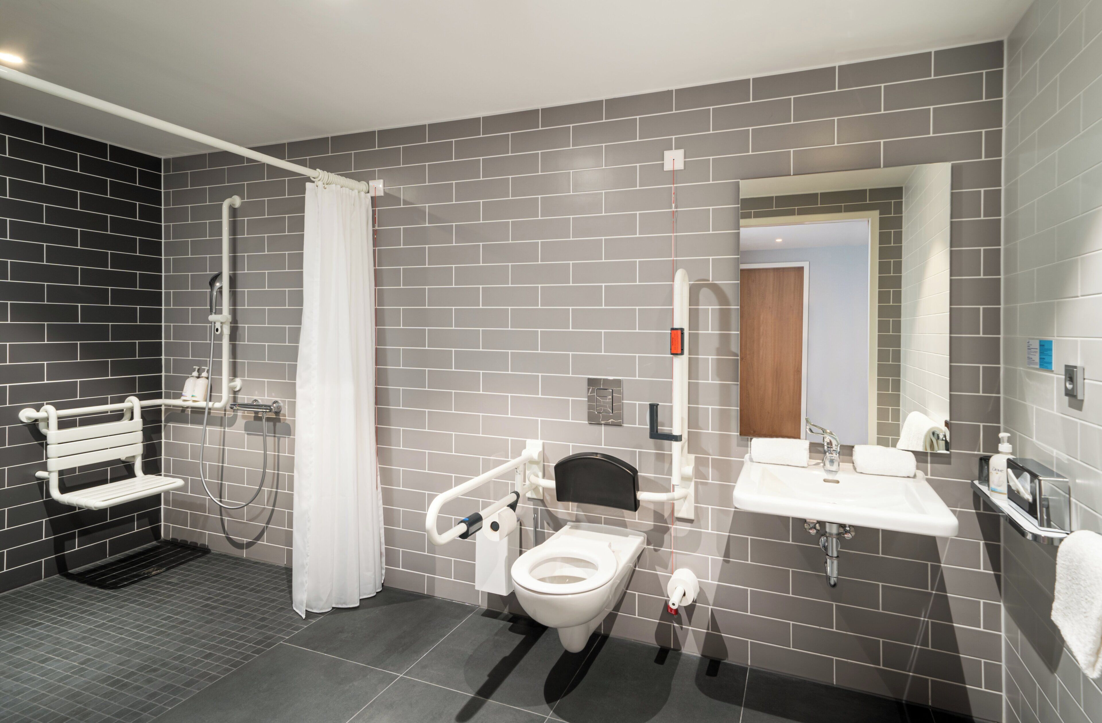 A modern bathroom with a toilet, bidet, and sink, featuring grey tiles and a white bench.