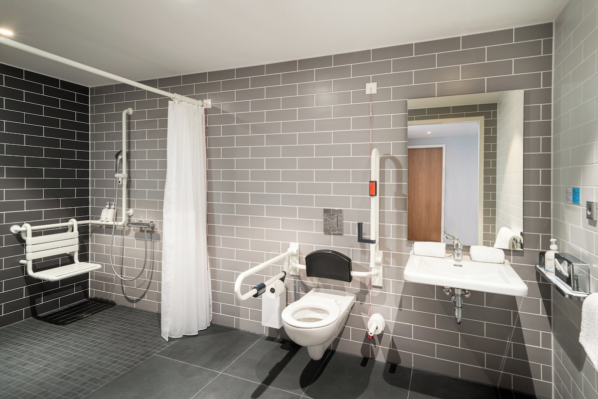 Un baño moderno con inodoro, bidé y lavamanos, con baldosas grises y un banco blanco.