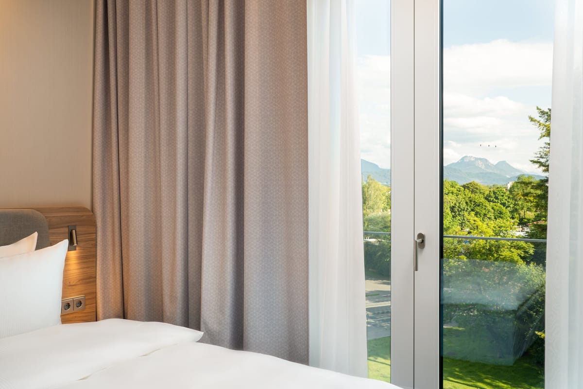Ein Hotelzimmer mit Bett, Vorhängen und Blick auf Berge durch eine Glastür.