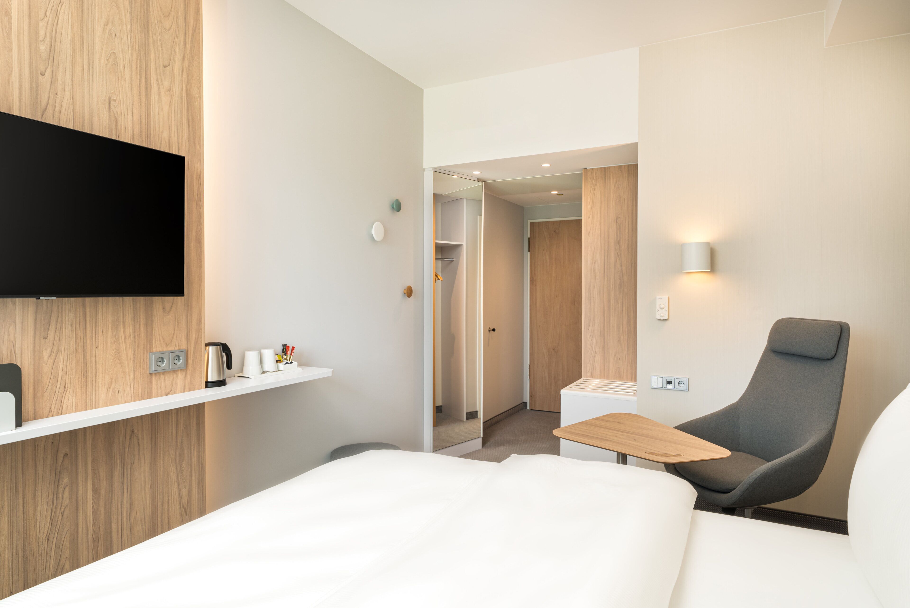 Una habitación de hotel moderna con una televisión de pantalla plana montada en la pared, un panel de madera decorativo, una zona de estar con una silla y una mesita, y un armario con espejo.
