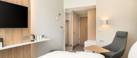 A modern hotel room with a flat-screen TV mounted on the wall, a wooden accent wall, a seating area with a chair and a small table, and a closet with a mirror.