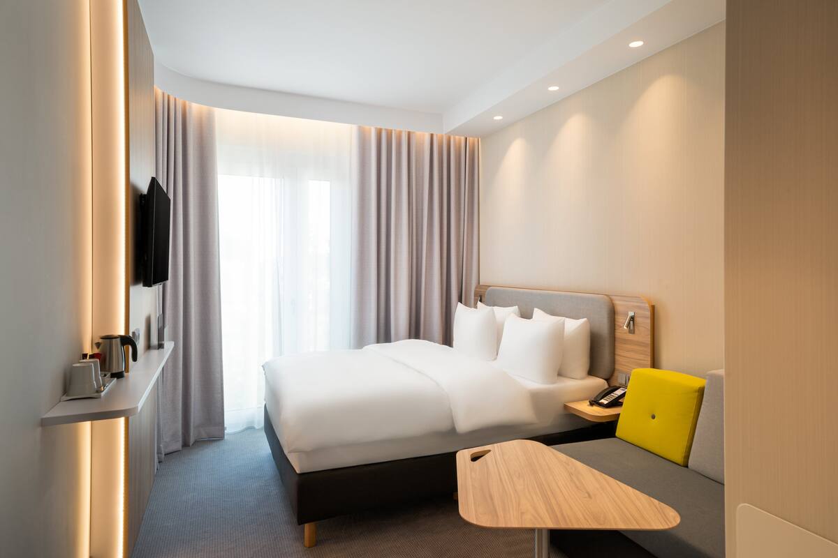 A modern hotel room with a large bed, bedside table, and a small desk with a chair.
