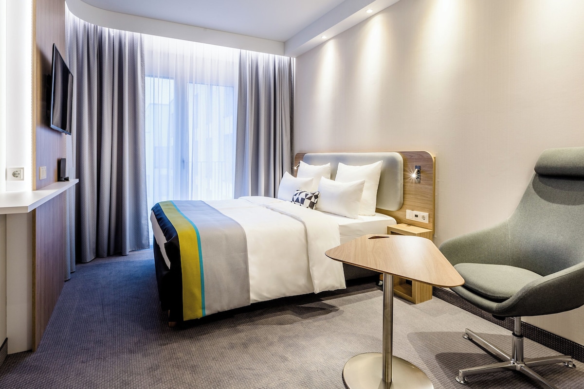 Habitación de hotel con cama, televisión montada en la pared, una silla, una mesita y una ventana con cortinas.