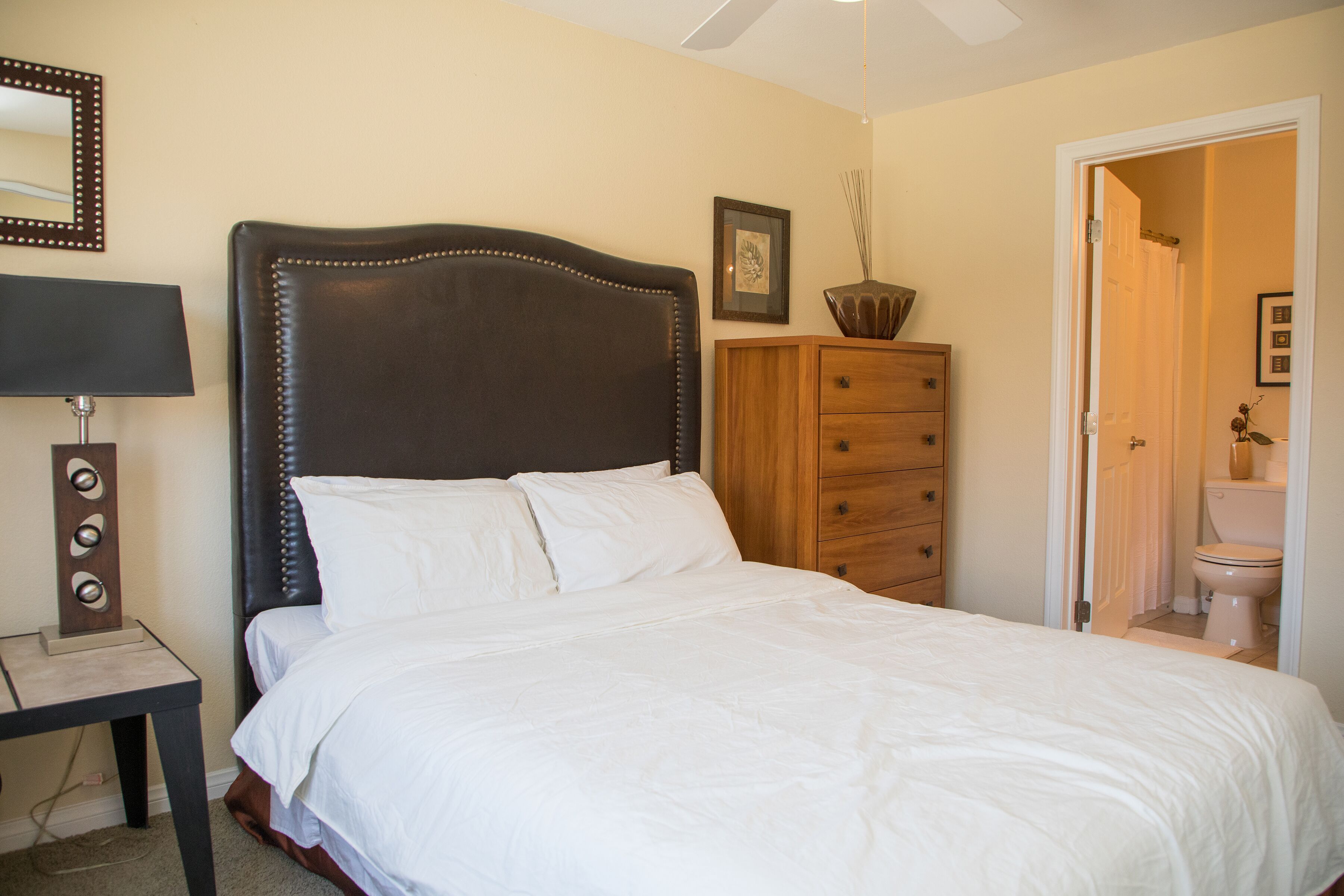A bedroom with a large bed, a wooden dresser, a nightstand with a lamp, and a door leading to a bathroom.