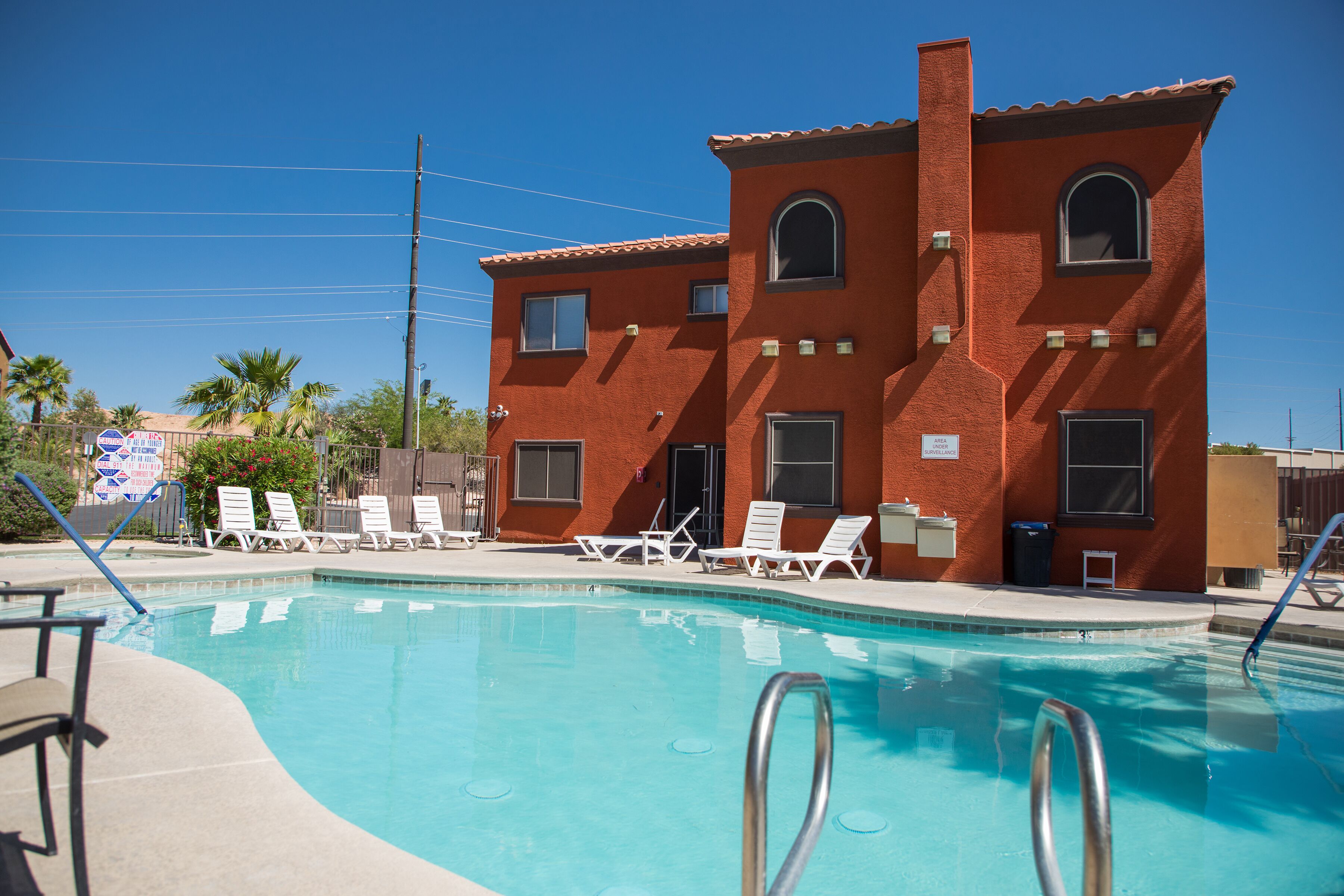 A red brick building with a swimming pool, white lounge chairs, and a clear blue sky.