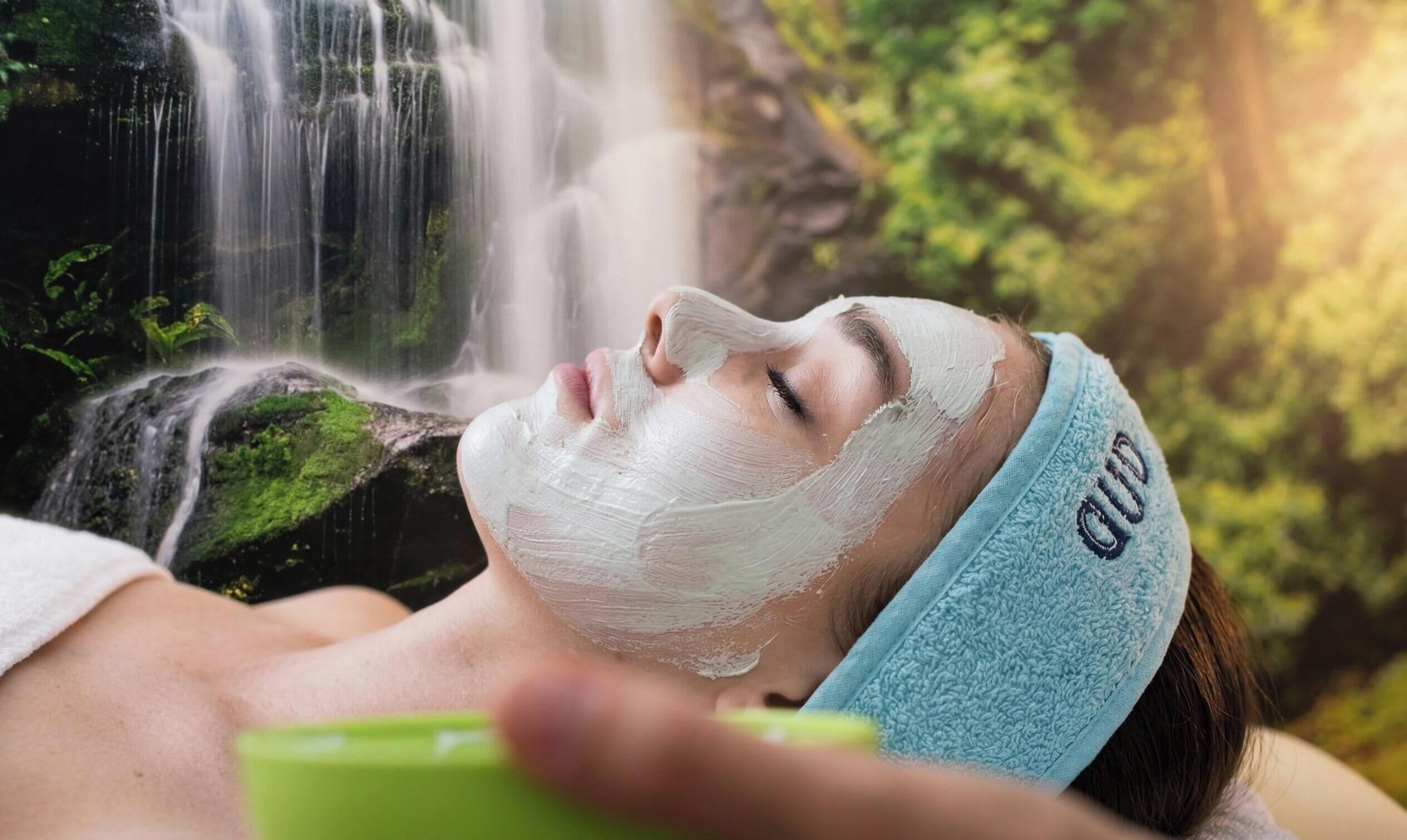 A person with a headband, lying down with a white substance on their face, in front of a waterfall.