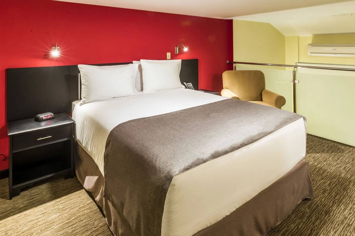 A hotel room with a large bed, a nightstand with a phone, a chair, and a red wall.