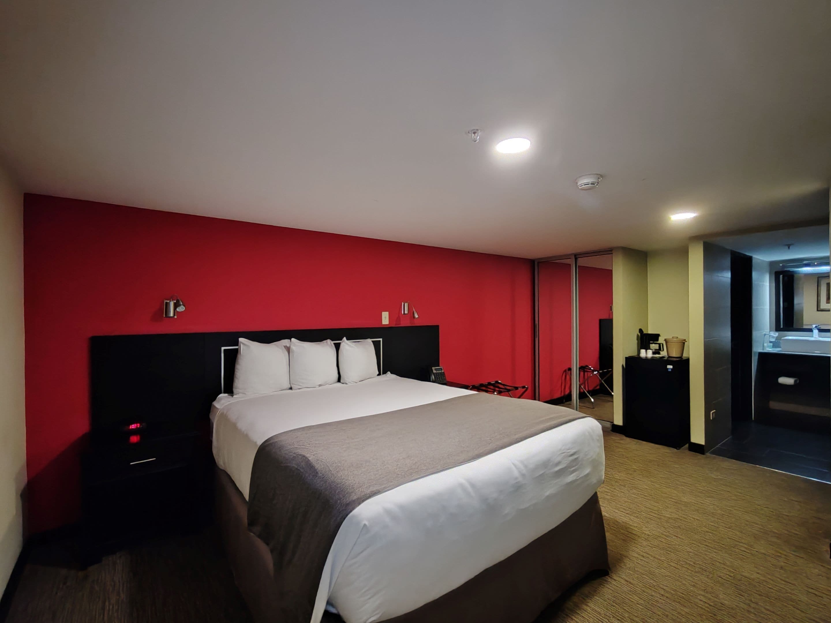 A hotel room with a large bed, a red accent wall, a small desk with a lamp, and a bathroom visible through a sliding door.