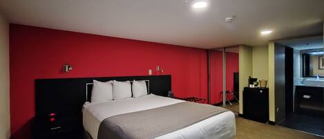 A hotel room with a large bed, a red accent wall, a small desk with a lamp, and a bathroom visible through a sliding door.