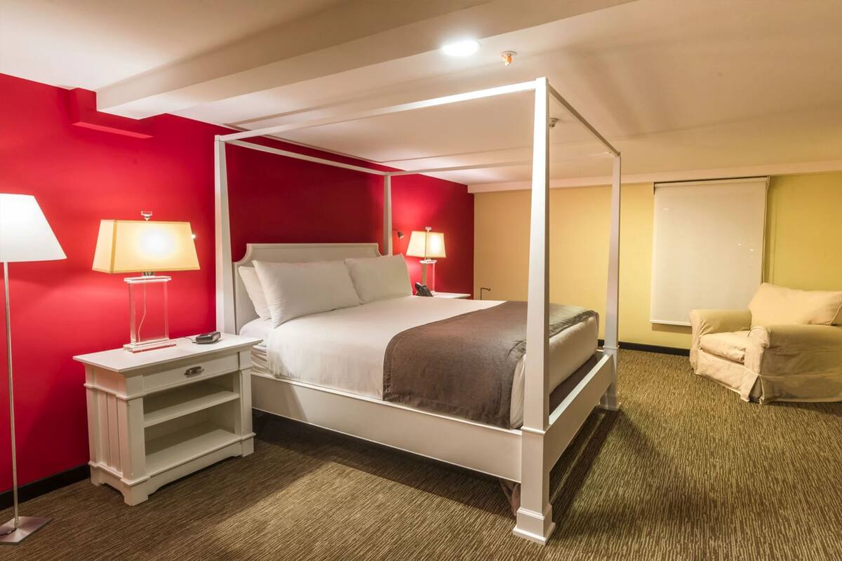 A four-poster bed with white bedding, two bedside tables with lamps, a beige armchair, and a large window with blinds.