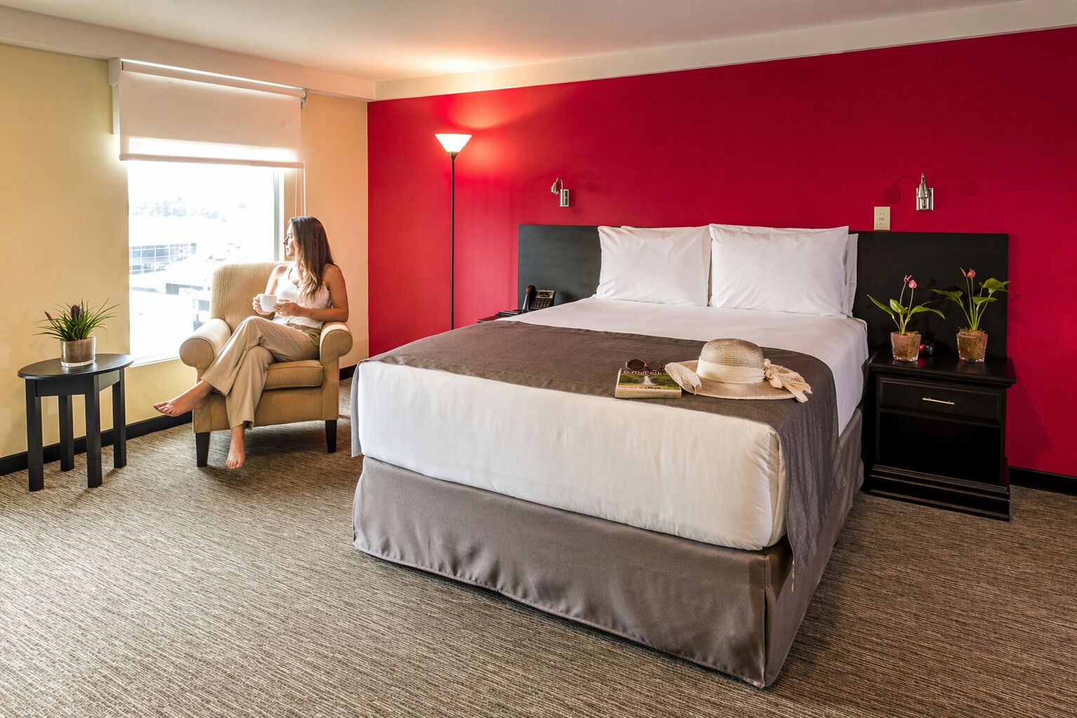 A hotel room with a bed, a chair, a nightstand, a lamp, and a window.