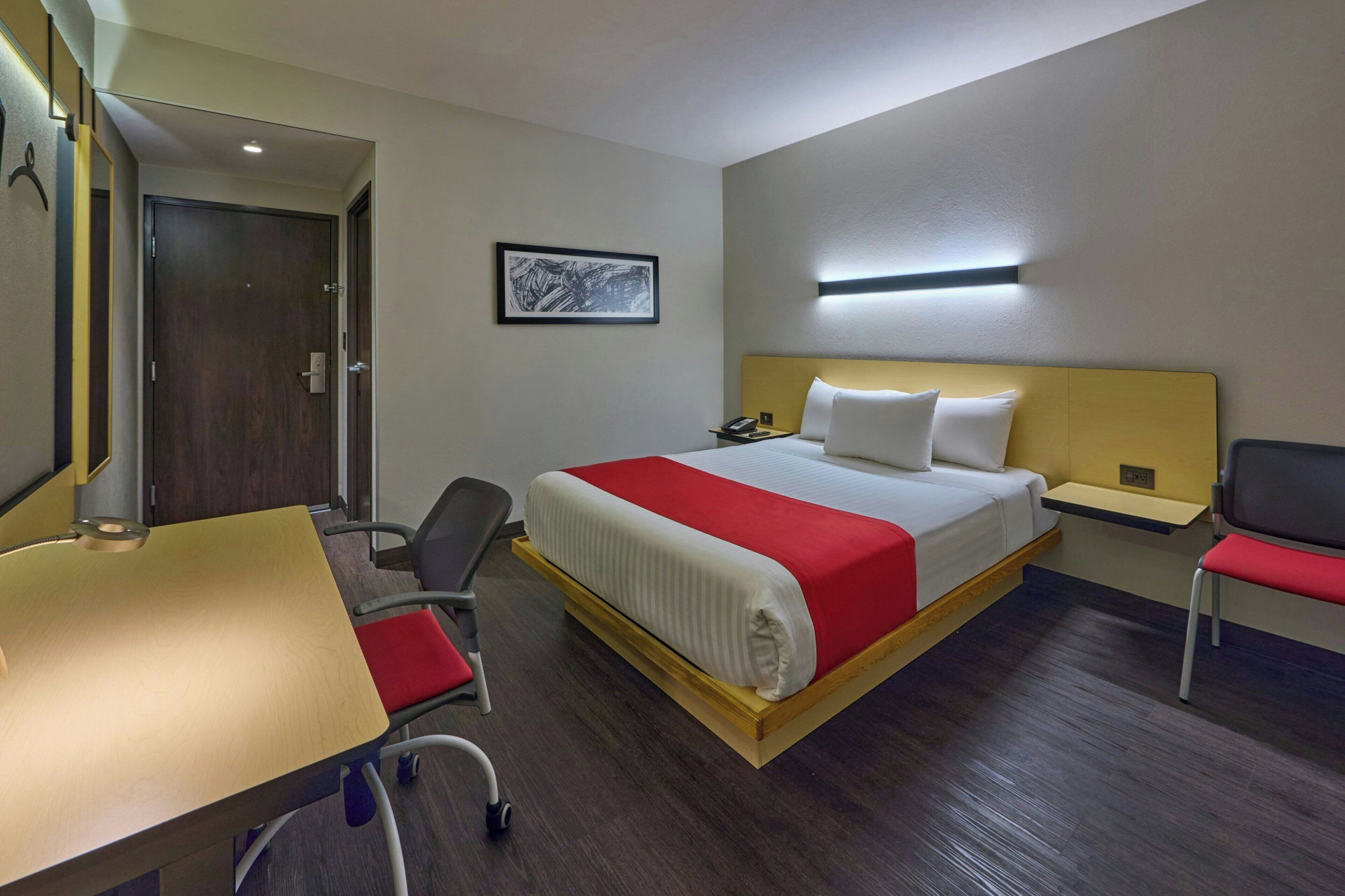Una habitación de hotel con cama, escritorio, silla, mesita de noche y puerta.