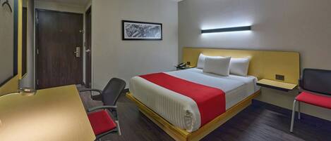 Una habitación de hotel con cama, escritorio, silla, mesita de noche y puerta.