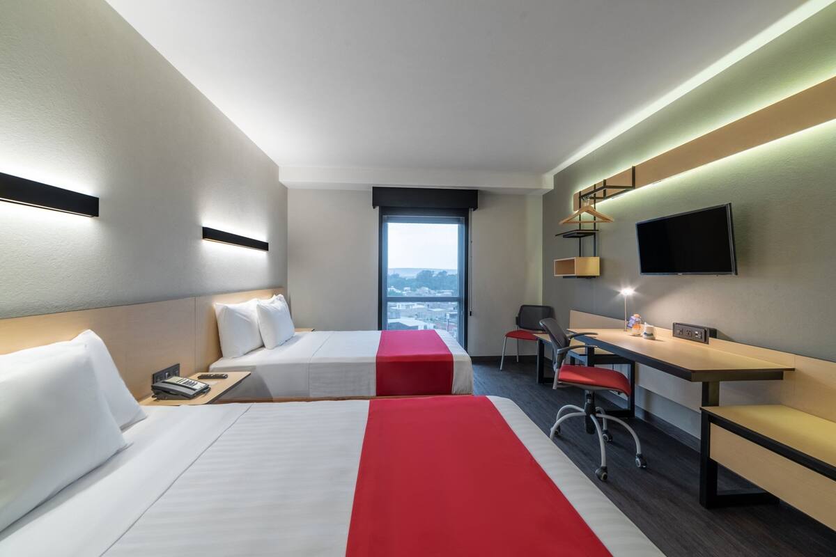 Habitació d'hotel amb dos llits, una taula de treball, una cadira, un televisor i una finestra amb vista a la ciutat.