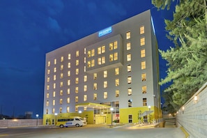 Un hotel de varios pisos con un cartel llamativo de 'City Express', un acceso cubierto y un área de estacionamiento con una camioneta blanca.