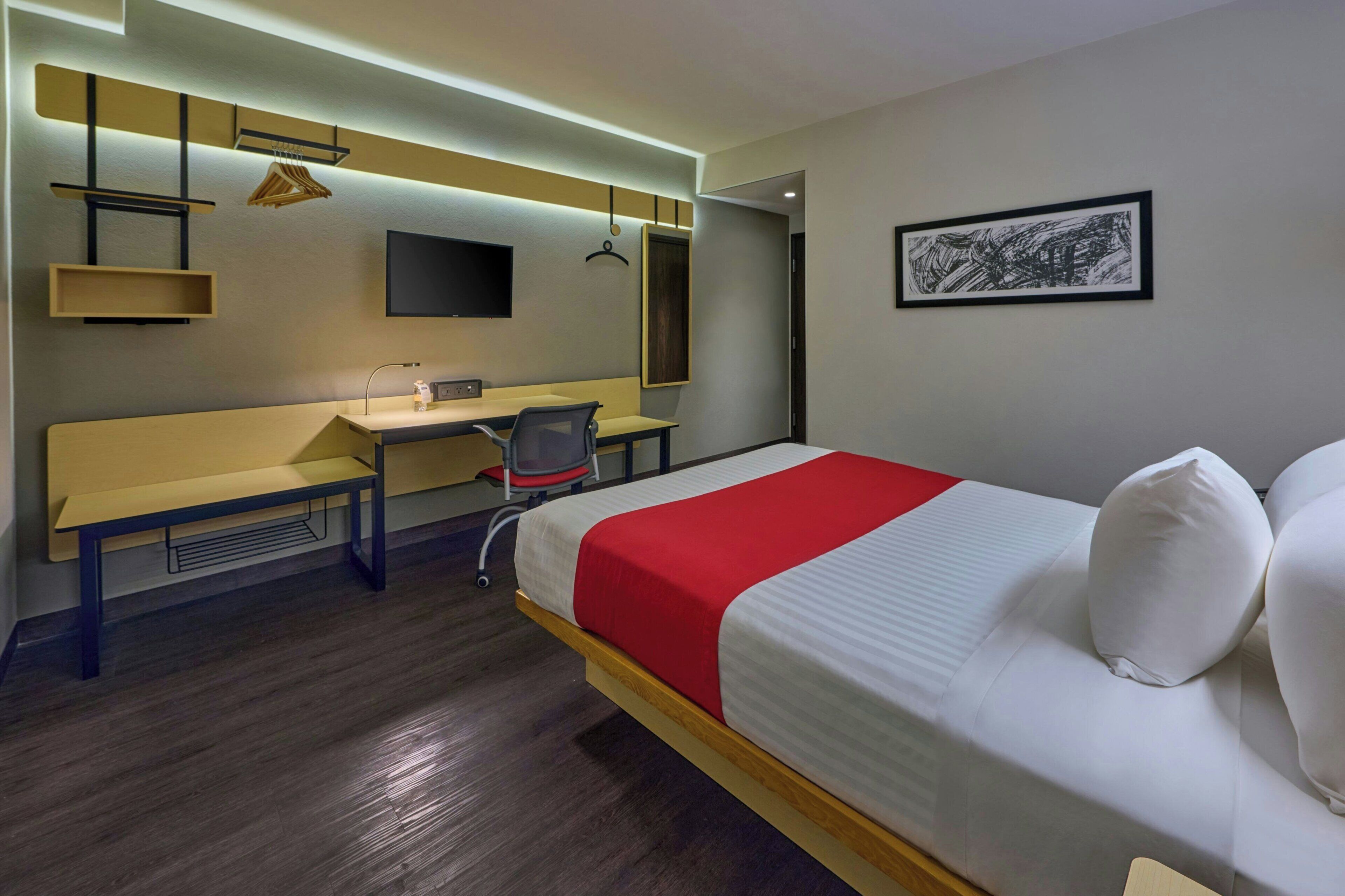 A hotel room with a bed, a desk, a chair, a TV, and a framed artwork on the wall.