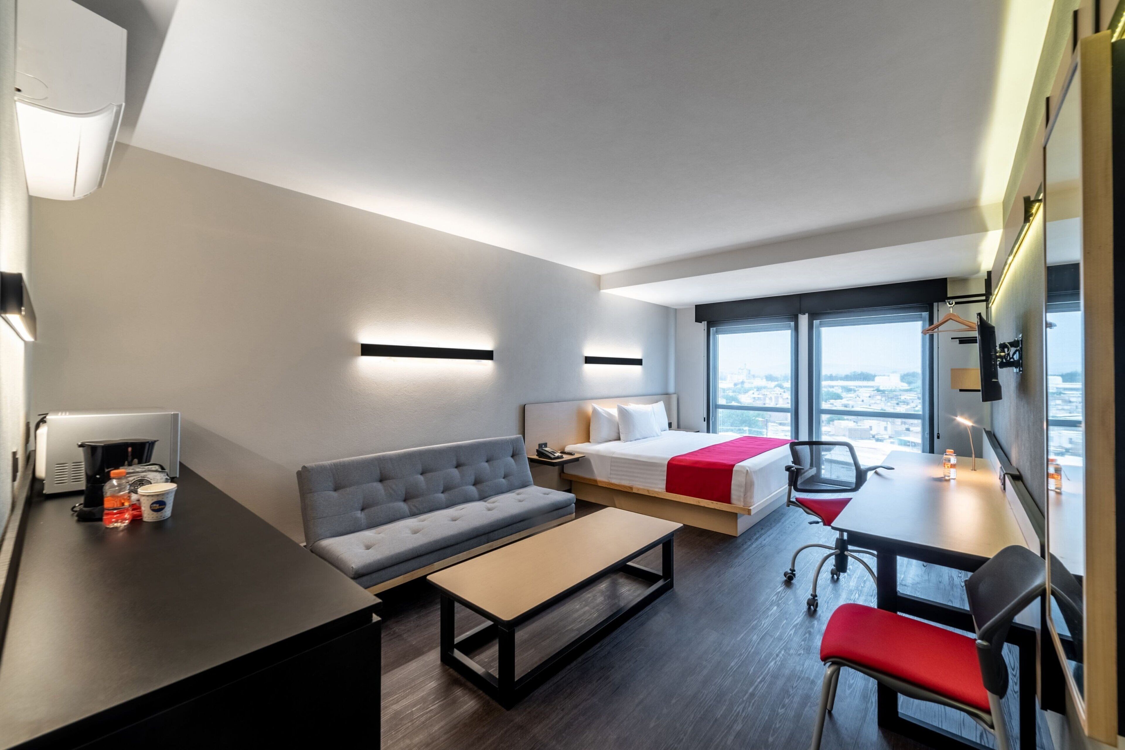 Una habitación de hotel moderna con sofá, cama, escritorio y ventanas amplias con vista a la ciudad.