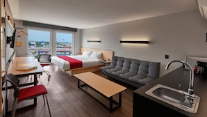 A modern hotel room with a bed, sofa, dining table, and kitchenette.