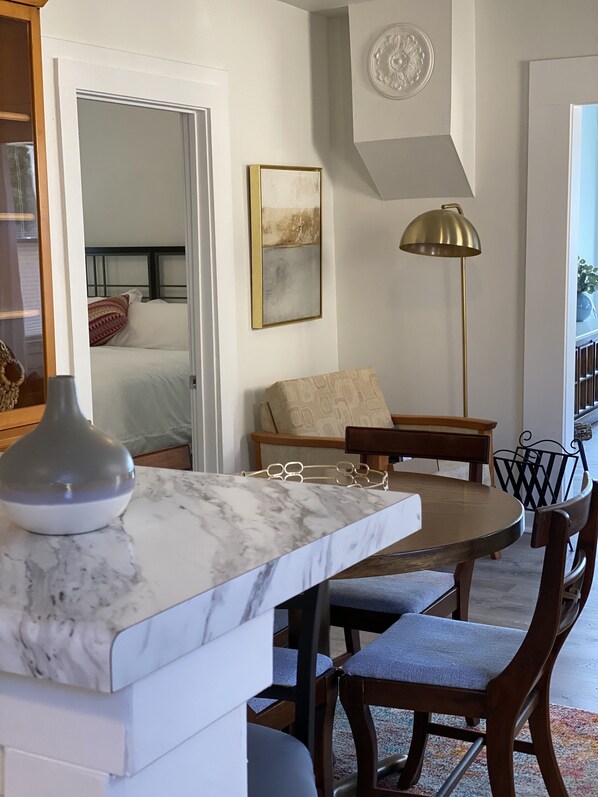 A modern kitchen with a marble countertop, a dining area with wooden chairs, a floor lamp, and a painting on the wall.