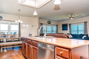 A modern kitchen with a central island, wooden cabinets, a dishwasher, pendant lights, a ceiling fan, a dining area with chairs, and a living area with a sofa.