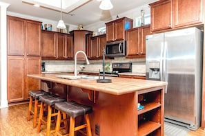 Una cocina con gabinetes de madera, un refrigerador de acero inoxidable, un microondas, una estufa y un lavamanos con un grifo moderno. Hay tres taburetes de bar en la isla.