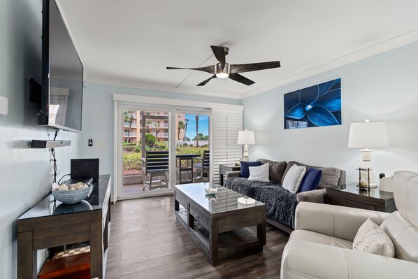 A modern living room with a sofa, coffee table, and a TV on a stand. There is a ceiling fan, a window with blinds, and a view of a balcony with outdoor furniture.