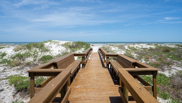 Een houten vlonder die naar een strand loopt, met duinen en een heldere lucht.