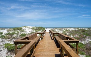 A wooden boardwalk leading to a beach with dunes and a clear sky.