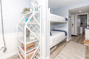A bunk bed with a white metal ladder, a small shelf with decorative items, and a rug on the floor. - Beachfront Bliss - Oceanfront 1 BR/1.5 BA at The Surf Condos! (Surf City)