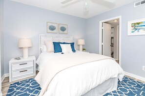 Une chambre à coucher comprenant un lit, des tables de chevet, une commode blanche, un tapis à motifs bleus et deux œuvres d’art murales.