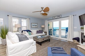 A living room with a white sofa, a coffee table, and a ceiling fan. There is a sign on the wall that reads 'EVERY SUMMER HAS A STORY.' - Beachfront Bliss - Oceanfront 1 BR/1.5 BA at The Surf Condos! (Surf City)