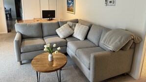 A grey sectional sofa with multiple cushions, a small wooden coffee table with a vase of white flowers, a flat-screen TV on a wooden stand, and a framed picture on the wall.