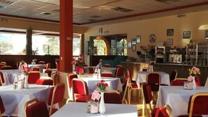 Um restaurante com várias mesas e cadeiras, cada uma com toalha branca e cadeiras vermelhas. Há arranjos florais sobre as mesas, e o restaurante possui um balcão com diversos itens expostos.