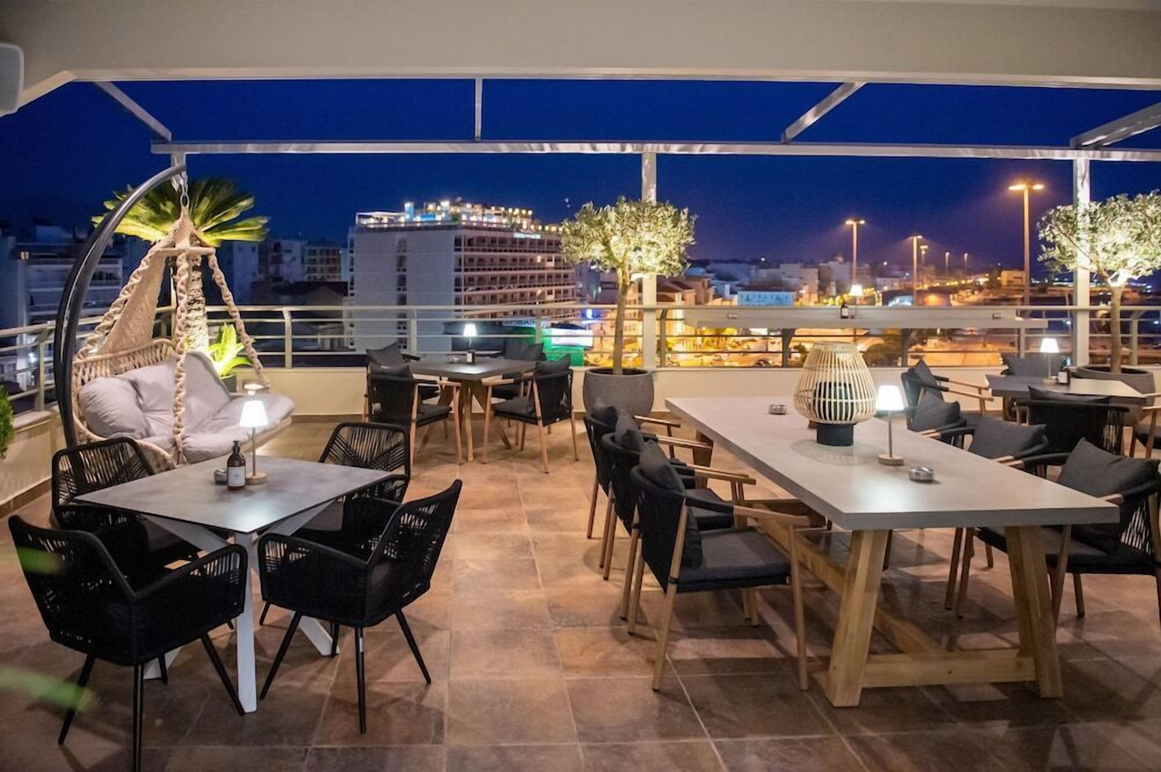 An outdoor dining area with tables and chairs, a hanging chair, and a view of the city at night.