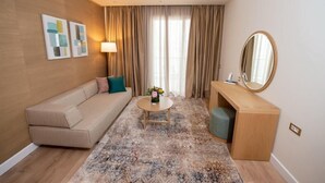 A hotel room with a sofa, a small round table with a vase, a desk with a mirror, and a large window with curtains. - Patras Palace Hotel (Patras)